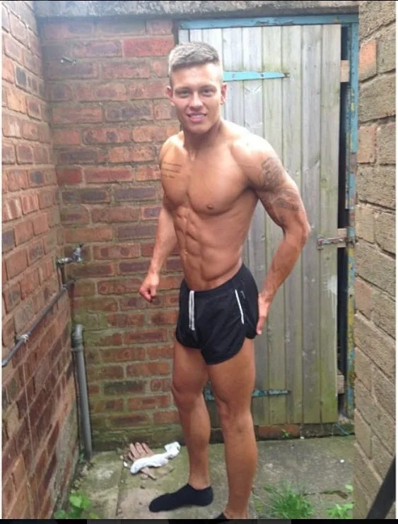 Muscular lad in tiny black shorts flexes abs against brick wall, one sock on, cheeky grin