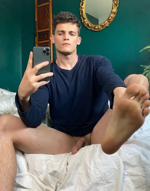 Hot guy takes a mirror selfie, bare feet up, cock peeking from under shirt