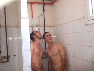 Two wet lads laughing under shower spray, bare chests glistening in tiled stall