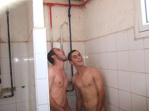 Two wet lads laughing under shower spray, bare chests glistening in tiled stall