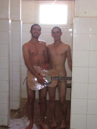 Two shirtless lads grinning in a tiled shower, one holding a guitar over his junk