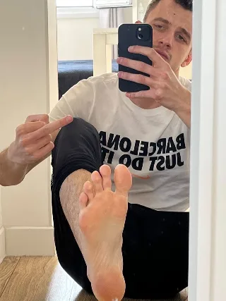 Guy in Nike tee points at his bare foot while snapping a mirror selfie, legs spread wide