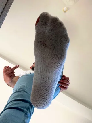 Guy in blue jeans and white tee teases with socked foot held high, thumb up, low-angle shot
