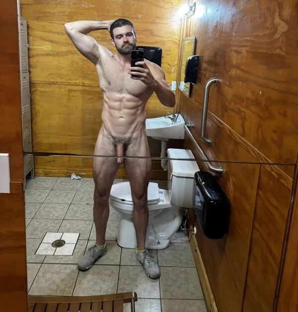 Hombre barbudo del gimnasio se toma selfie frente al espejo mostrando su polla desnuda y erección dura en un cubículo de baño