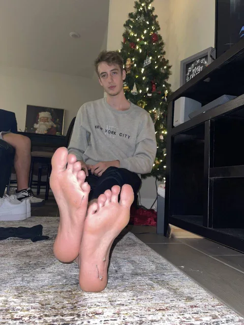 Young lad in grey sweatshirt shows off bare soles with Christmas tree behind him