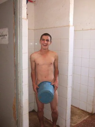 Smiling naked lad holds blue bucket over his cock in tiled shower stall