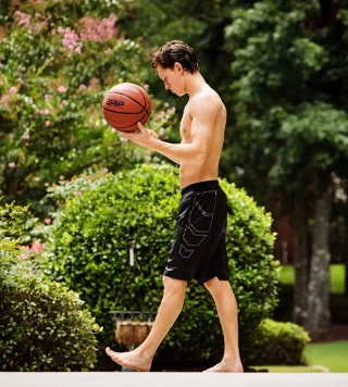 Tom Holland shirtless with basketball, barefoot on court, lean abs and athletic shorts in garden light