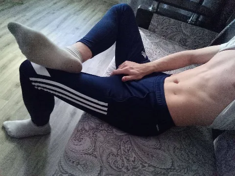 Shirtless lad in Adidas track pants flexes socked feet while lounging bare-chested on patterned couch