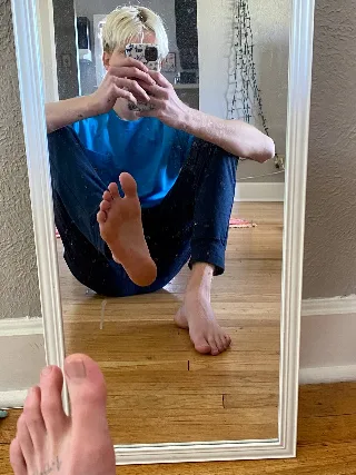 Blonde guy in blue tee mirrors his bare feet, one lifted high for foot-fetish focus