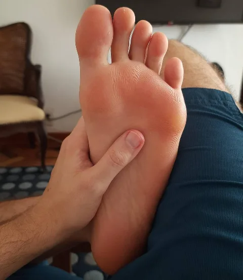 Guy gently massaging his own bare foot, blue shorts on, soles up close