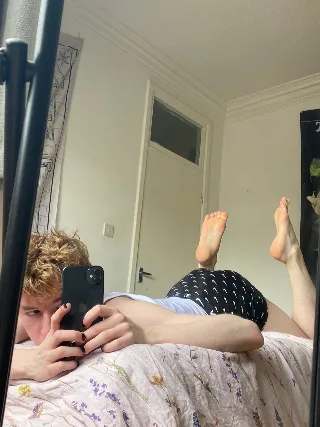Curly-haired lad snaps mirror selfie with bare feet up, shorts riding high on bed