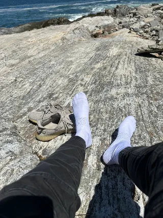 Guy in white socks and black pants lounging barefoot on coastal rocks with sneakers nearby