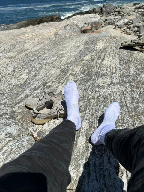Guy in white socks and black pants lounging barefoot on coastal rocks with sneakers nearby