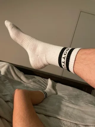 Lazy lad in white athletic socks flexing feet on rumpled sheets, one leg kicked high