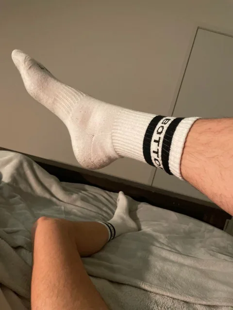 Lazy lad in white athletic socks flexing feet on rumpled sheets, one leg kicked high