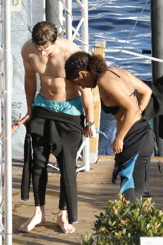 Tom Holland shirtless on dock, barefoot and lean, pulling on wetsuit over turquoise swim trunks