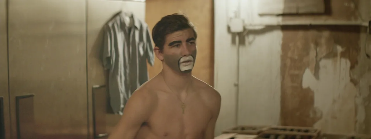 Clown-nosed celeb in tank top poses against gold fringe, shirtless moments captured mid-undress in gritty room