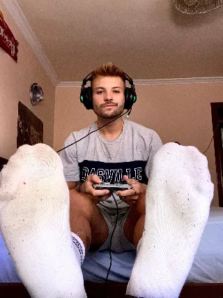 Guy in gaming headset flexes socked feet toward camera while holding controller, shorts riding high