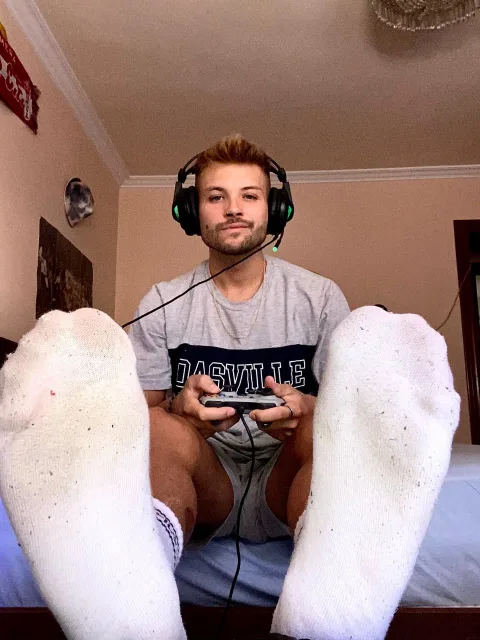 Guy in gaming headset flexes socked feet toward camera while holding controller, shorts riding high