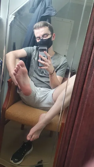Guy in grey tee and shorts snaps mirror selfie with bare feet up, one sole pressed to thigh