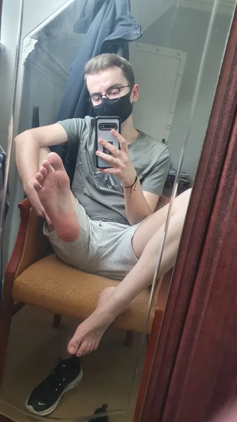 Guy in grey tee and shorts snaps mirror selfie with bare feet up, one sole pressed to thigh