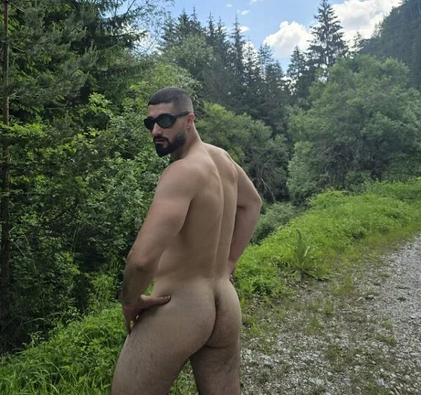 Cara musculoso com barba e óculos escuros exibe sua bunda pelada ao ar livre, mão no quadril