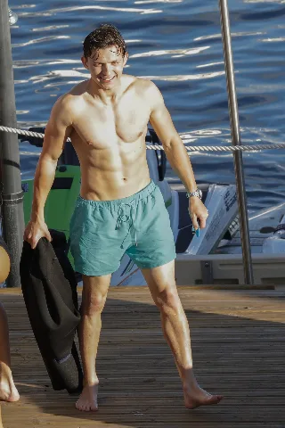 Tom Holland shirtless on deck, smiling with sea behind, holding wetsuit, mint shorts hugging his lean frame
