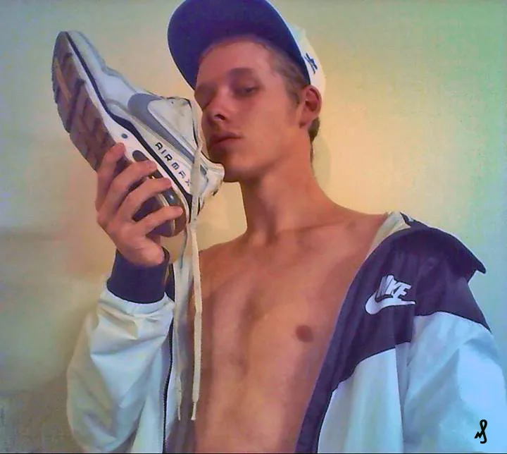 Garoto sem camisa de jaqueta Nike e boné segurando tênis em seus lábios